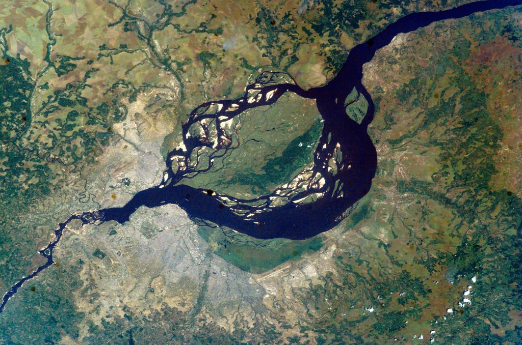 A view of the twin Congo capitals, facing each other across the Congo River. In this 2003 photo taken from the International Space Station, the smaller city of Brazzaville, capital of the Republic of the Congo, is at upper left. The much larger grey area at lower left is Kinshasa, capital of the Democratic Republic of the Congo. The cities lie at the point where the Congo River becomes navigable upstream, widening to the east into Pool Malebo – previously named Stanley Pool by the brutal 19th century British-American explorer Henry Morton Stanley, who prospected the region on behalf of King Leopold II of Belgium.