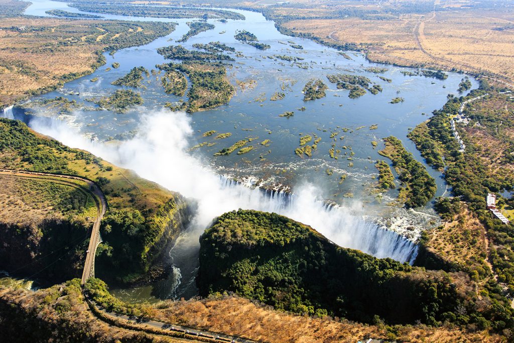 An aerial view of the massive Zambezi River – which gives Zambia its name – and the Victoria Falls. The waterfall, named Mosi-oa-Tunya or “the smoke that thunders” in Tokaleya Tonga, is said to be the biggest in the world. Its combined width of 1,708 metres and height of 108 metres result in the world’s largest sheet of falling water. Here Zambia’s border with Zimbabwe runs along the middle of the river, with Zimbabwe on the left and Zambia on the right. The steel Victoria Falls bridge at lower left is a border post between the countries.
